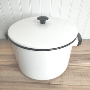 May include: A white enamel pot with a black rim and handles. The pot has a matching lid with a black knob. The pot is sitting on a wooden surface.