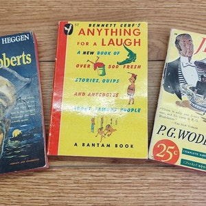 May include: Three vintage paperback books with colorful covers. The book on the left is titled "Mister Roberts" and features a man in a navy uniform. The middle book is titled "Anything for a Laugh" and features a cartoon illustration of a man in a green swimsuit. The book on the right is titled "Jeeves" and features a man in a tuxedo.