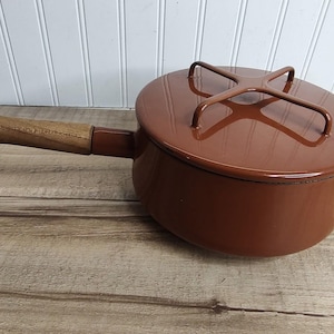 May include: A round, brown enameled metal saucepan with a matching lid featuring a cross-shaped handle. The pan has a long, straight handle with a light-colored wooden grip. The exterior finish is smooth and glossy, reflecting light on the surface.