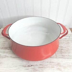May include: A red enamel cast iron pot with white enamel interior and two handles. The pot is empty and sitting on a white wooden surface.