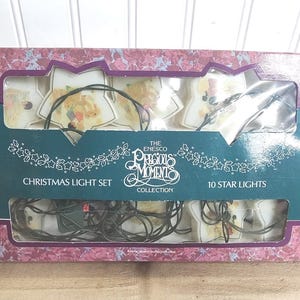 May include: A boxed Christmas light set featuring ten star-shaped lights. The packaging is a mix of pink and purple with a teal label that reads "Precious Moments Collection" and "Christmas Light Set".