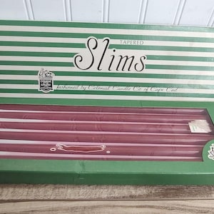 May include: A box of Colonial Candle Co. of Cape Cod Tapered Slims candles. The packaging is green and white striped with the text Slims and Colonial Candle Co. of Cape Cod. Inside the box are several long, thin, dark red tapered candles.