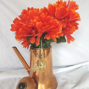 May include: A copper teapot with a clock face and hands, used as a vase for a bouquet of orange flowers.