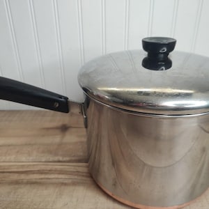 May include: A stainless steel saucepan with a copper base, featuring a black handle with a metal hanging loop and a matching stainless steel lid with a black knob. The pot has a cylindrical body and a polished metallic finish throughout.