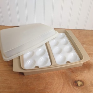 May include: A beige plastic egg tray with a matching lid. The tray has two white inserts, each with six egg-shaped indentations. The lid is partially open, revealing the interior. The tray is designed for storing eggs.