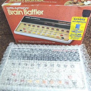 May include: Vintage Mattel Electronics Brain Baffler game in its original box. The box is red with the game's name and logo. The game console is white and black with multiple buttons. The game is wrapped in bubble wrap.