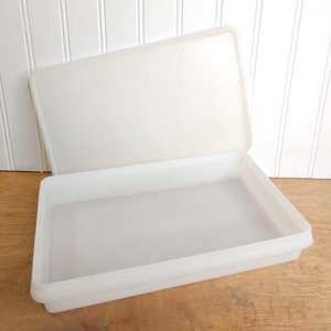 May include: A rectangular, translucent white plastic container with a matching lid. The lid is slightly ajar, revealing the interior of the container. The container is sitting on a wooden surface, with a white paneled wall in the background.