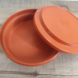 May include: An open, round, orange-colored container with a matching lid. The lid is slightly ajar, revealing the interior of the container. The container and lid are made of a solid, durable material, likely plastic. The design is simple and functional.