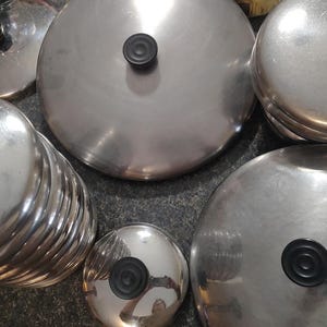 May include: A stack of silver-colored metal pot lids with black handles. The lids are arranged in a pile, with some overlapping. The lids are all the same size and shape.