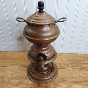 May include: Antique copper tea or coffee dispenser with a spigot and a lid with a black knob. The dispenser has a round base and two handles.