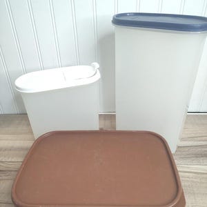 May include: Three plastic food storage containers. Two are tall and rectangular with white bodies and colored lids. The third is a rectangular container with a brown lid. All containers are empty and ready for use.