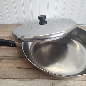 May include: A stainless steel skillet with a polished finish, featuring a long black handle and a matching circular lid with a black knob. The pan has a wide, shallow basin and a smooth interior surface, designed for stovetop cooking and heat retention.