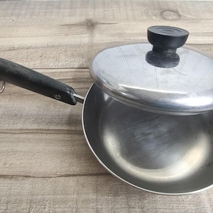 May include: A stainless steel skillet with a black handle and a matching lid. The handle features a small metal hanging loop at the end. The lid has a black knob handle in the center. The pan has a polished interior and a smooth, rounded rim.