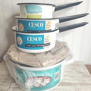 May include: A stack of vintage white enamel cookware with black handles. The set includes three saucepans and a larger cooking pot. The saucepans have blue and white labels with the brand name "CESCO". The cooking pot has a blue and white label that says "Cooking Pot".