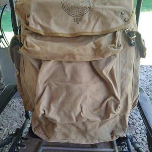 May include: A tan canvas backpack with a brown leather strap and a Boy Scouts of America patch with the text "Be Prepared" and "1307".