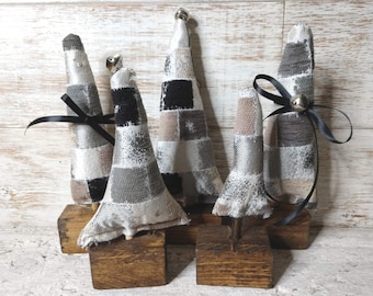 May include: Four small, handmade, fabric Christmas trees with a patchwork design in shades of gray, black, and white. Each tree is decorated with a black ribbon and a silver bell. The trees are displayed on wooden bases.