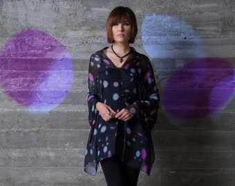 Op de afbeelding: Een vrouw draagt een zwart-wit geblokt shirt met een transparante overlay. Het shirt heeft een knoopsluiting aan de voorkant en lange mouwen. Ze draagt een zwarte legging en zwarte hakken. De achtergrond is een grijze betonnen muur met paarse en blauwe cirkels.