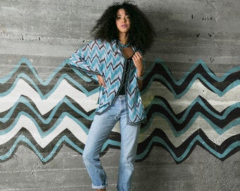 Op de afbeelding: Een vrouw draagt een blauw-wit gestreept shirt met een zwart onderhemd, een blauwe jeans en zwarte enkellaarsjes. Ze staat voor een muur met een zwart-wit chevronpatroon.