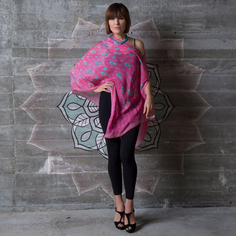 Op de afbeelding: Een vrouw draagt een roze en blauwe sjaal met een zwart topje en zwarte leggings. Ze staat voor een betonnen muur met een geschilderd mandala-ontwerp.
