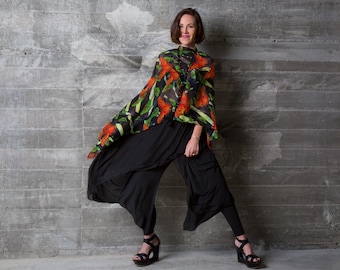 Op de afbeelding: Een vrouw draagt een zwarte outfit met een kleurrijke top met bloemenpatroon. De top heeft een mix van oranje, groene en zwarte kleuren. De outfit bestaat uit een zwarte broek en zwarte plateausandalen. De achtergrond is een grijze betonnen muur.