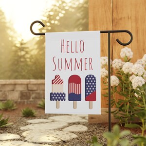 May include: A white garden flag with the words "HELLO SUMMER" in red. The flag features three popsicle designs with red, white, and blue patterns. The flag is displayed on a black metal stand in a garden setting.