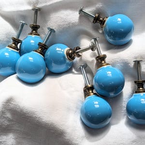 May include: A collection of eight light blue ceramic cabinet knobs with a glossy finish. Each knob has a brass-colored base and a silver screw for attachment. The knobs are arranged on a white fabric surface.