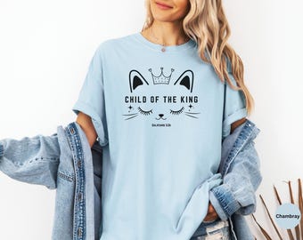 May include: Light blue t-shirt with a black graphic of a cat face wearing a crown, with the text "CHILD OF THE KING" and "GALATIANS 3:26". The shirt has short sleeves and a relaxed fit.