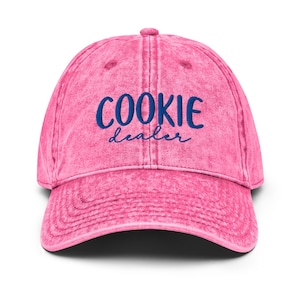 May include: A pink, distressed baseball cap with the words "COOKIE dealer" embroidered in blue script across the front. The cap has a curved brim and a button on top. The hat is a vintage style.