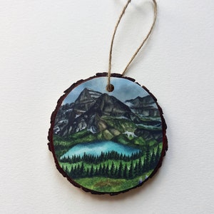 May include: A round wooden ornament with a landscape painting of mountains, a lake, and evergreen trees. The ornament is approximately 3 inches in diameter and has a natural bark edge. A twine loop is attached for hanging.