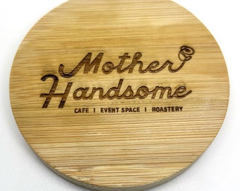 May include: A round, wooden coaster with the text "Mother Handsome" in a cursive font. Below the text, it reads "Cafe | Event Space | Roastery".
