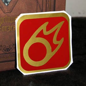 Peut inclure: Un livre relié en cuir marron avec des accents dorés et un motif décoratif. Le livre a un grand cristal transparent incrusté dans la couverture. Le titre "Encyclopedia Eorzea" est visible sur la couverture. Un autocollant rouge et or avec un motif de flamme stylisé est collé sur le livre.
