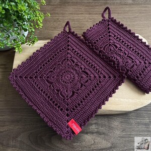Könnte beinhalten: Zwei quadratische, dunkelviolette gehäkelte Topflappen mit einem zentralen Blumenmuster und einem gewellten Rand. Jedes Stück hat eine kleine Aufhängeschlaufe an der oberen Ecke und ein rotes Stoffetikett mit dem weißen Schriftzug "Mäusey".