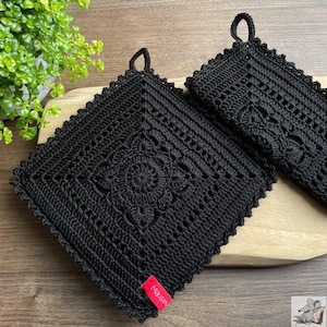 Könnte beinhalten: Zwei quadratische, schwarz gehäkelte Topflappen mit einem zentralen Blumenmuster und einem gewellten Rand. Jedes Stück hat eine kleine Schlaufe zum Aufhängen und ein rotes Stoffetikett mit dem weißen Text "mauseu" an der unteren Ecke.
