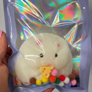 May include: A white, hamster-shaped bath bomb with black eyes and a yellow cheese wedge in its mouth, surrounded by colorful pom-poms. The bath bomb is packaged in a clear, iridescent pouch with a lavender zipper.