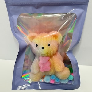 May include: A small, knitted teddy bear with a pink scarf, packaged in a clear, iridescent pouch inside a lavender-colored bag. The bear is yellow and white with black eyes. Colorful pom-poms are at the bear's feet.