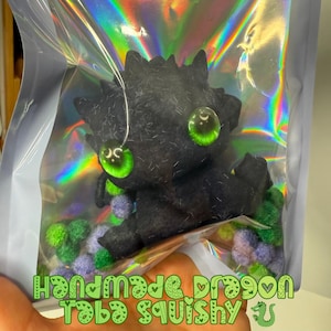 May include: A handmade black dragon plushie with large green eyes, displayed in a holographic bag. The plushie has a textured appearance and is surrounded by colorful pom-poms. Text on the bag reads "Handmade Dragon Toba Squishy".