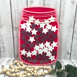 May include: A red mason jar-shaped decoration filled with white and glittery purple stars. The jar has a clear outline and a decorative rim. The background is a white wooden surface.