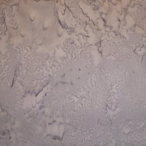 Op de afbeelding: Abstract textiel met een getextureerde, gevlekte uitstraling. De stof heeft een mix van lichtgrijze en gebroken witte tinten, met donkergrijze accenten die een patroon creëren dat doet denken aan een topografische kaart of een verweerd oppervlak.