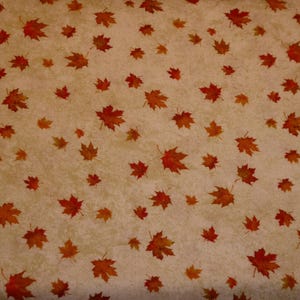 Op de afbeelding: Een stof met een herhaald patroon van oranje en rode esdoornbladeren verspreid over een beige achtergrond. De bladeren, van verschillende grootte, zijn gedetailleerd met aderen, wat een herfstthema creëert.