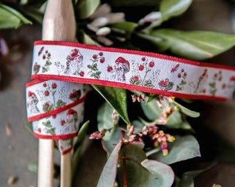 Pode incluir: Fita branca com borda vermelha, com um padrão repetido de cogumelos, bagas e folhagens. A fita é enrolada em um pino de madeira, com folhas verdes e pequenas flores ao fundo. O texto "acufactum Ute Menze" está na parte inferior.