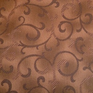 May include: Close-up of brown fabric featuring a repeating pattern of swirling, stylized floral designs. The fabric has a textured appearance with diagonal lines, creating a subtle grid effect. The colour palette is a mix of brown and darker brown.