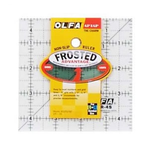 Può includere: Un righello in plastica trasparente con una griglia e un'etichetta gialla che dice "OLFA Frosted Advantage" con un logo verde e giallo. Il righello è quadrato di 11,4 cm.