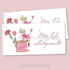 May include: Two white cards featuring a watercolour illustration of a fairy in a pink dress, holding a heart-shaped wand, and seated on a pink thread spool. Red flowers and the text "Mit Liebe selbstgemacht" are visible.