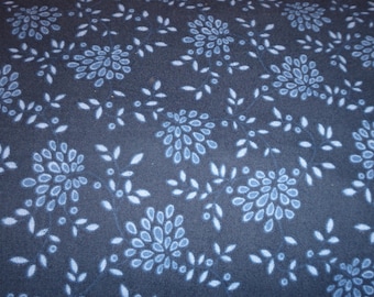 May include: Close-up of a fabric with a repeating floral pattern. The design features stylized blue flowers and white leaves on a dark blue background. The fabric has a textured appearance.