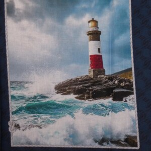 Può includere: Un quadrato di tessuto con una scena costiera di un faro. Il faro, rosso, bianco e nero, si erge su uno sperone roccioso, con le onde che si infrangono contro di esso. Il cielo è nuvoloso e il mare è turchese.