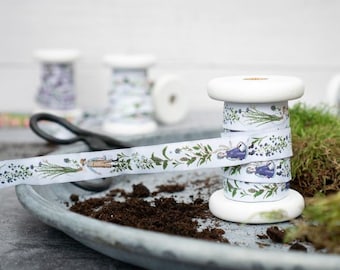 Pode incluir: Um carretel branco de fita com uma estampa floral e botânica que apresenta uma pequena figura com um vestido azul. A fita está em um carretel de madeira e está sentada em uma bandeja com terra.