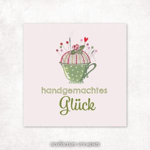 Könnte beinhalten: Eine Illustration zeigt eine grüne, gepunktete Teetasse mit einem Nadelkissen, das mit Nadeln und einem Herz gefüllt ist. Der Text "handgemachtes Glück" steht in grüner Schrift darunter. Der Name des Künstlers steht unten.