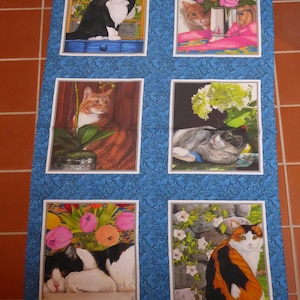 Op de afbeelding: Een stoffen paneel met een blauwe achtergrond en een witte rand. Het paneel toont zes verschillende afbeeldingen van katten in verschillende omgevingen. De katten worden afgebeeld in verschillende poses, waaronder slapen, zitten en staan. De afbeeldingen worden omringd door een decoratief bloemenpatroon.