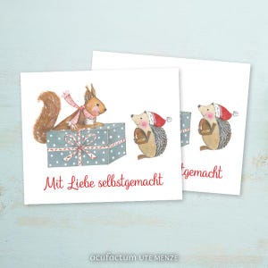May include: A white card with a red squirrel holding a gift box and a hedgehog with a red hat holding a small gift. The text on the card reads "Mit Liebe selbstgemacht".