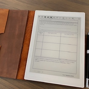 May include: A white e-reader with a brown leather case. The e-reader is displaying a page with a table and the text "1 to 1 Sessions 2024". A black pen is lying next to the e-reader.